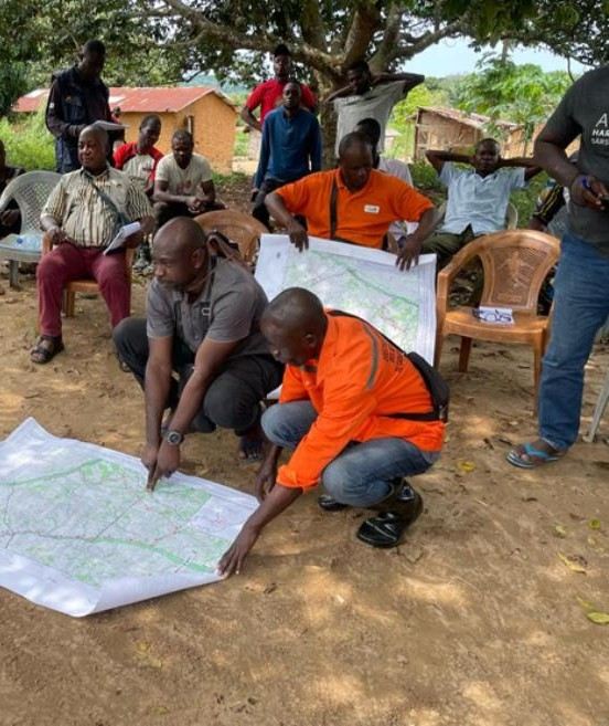 Land administration and project staff explain a map to residents seated under a tree in a village