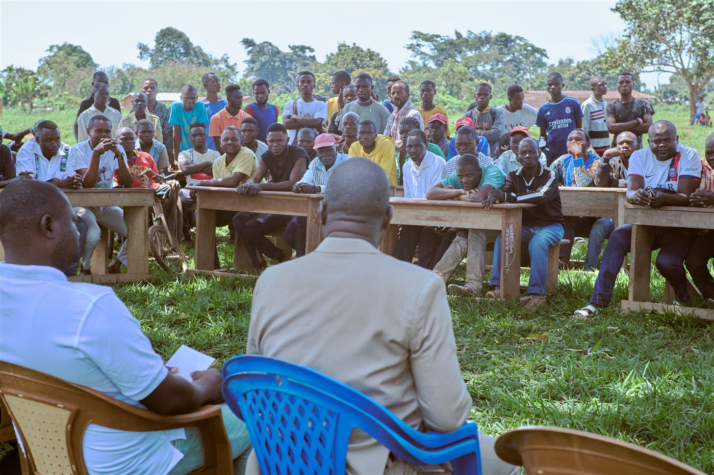 Residents seated at wooden benches under the trees face local officials at a village gathering